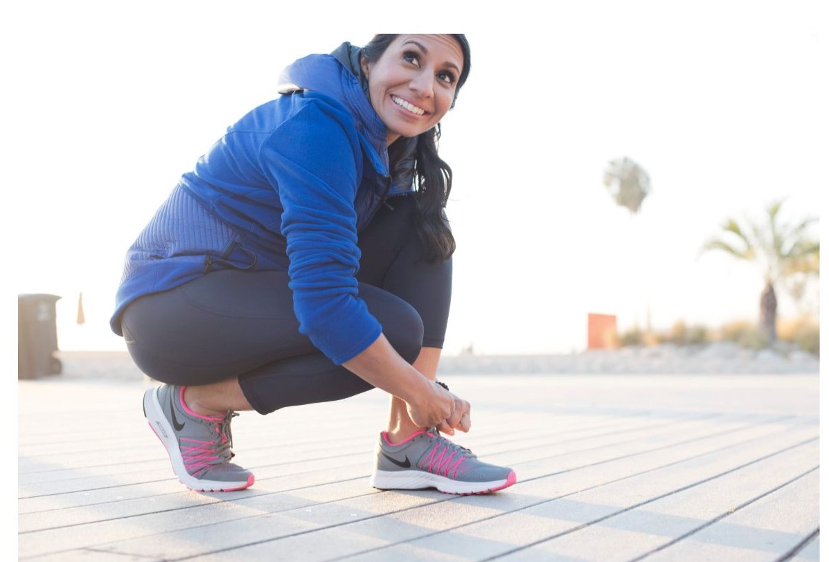 A woman in a blue jacket crouches on a wooden surface while tying the laces of her gray and pink sneakers.