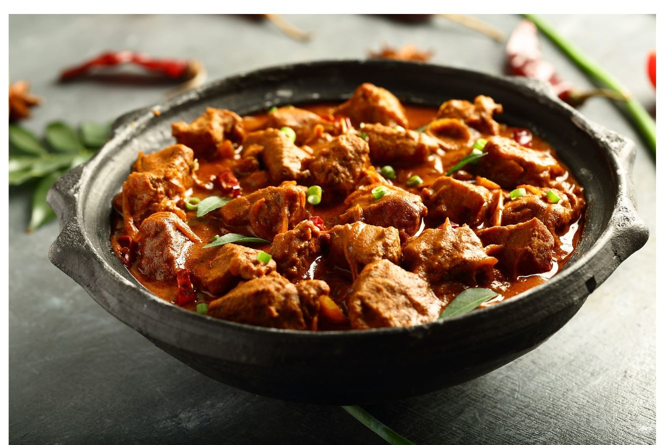 A clay pot filled with pieces of meat in a rich, reddish-brown sauce, garnished with green onions and curry leaves.