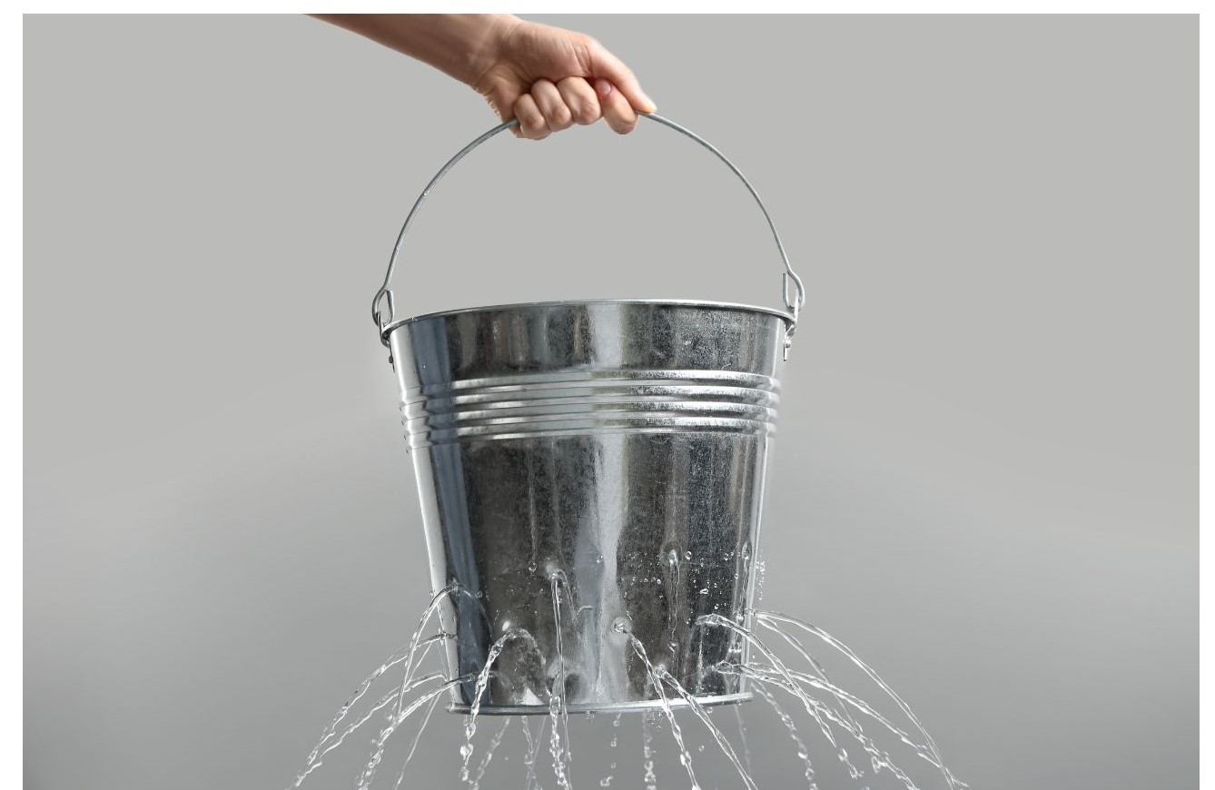 A hand holds a metal bucket pouring water from multiple streams against a gray background.