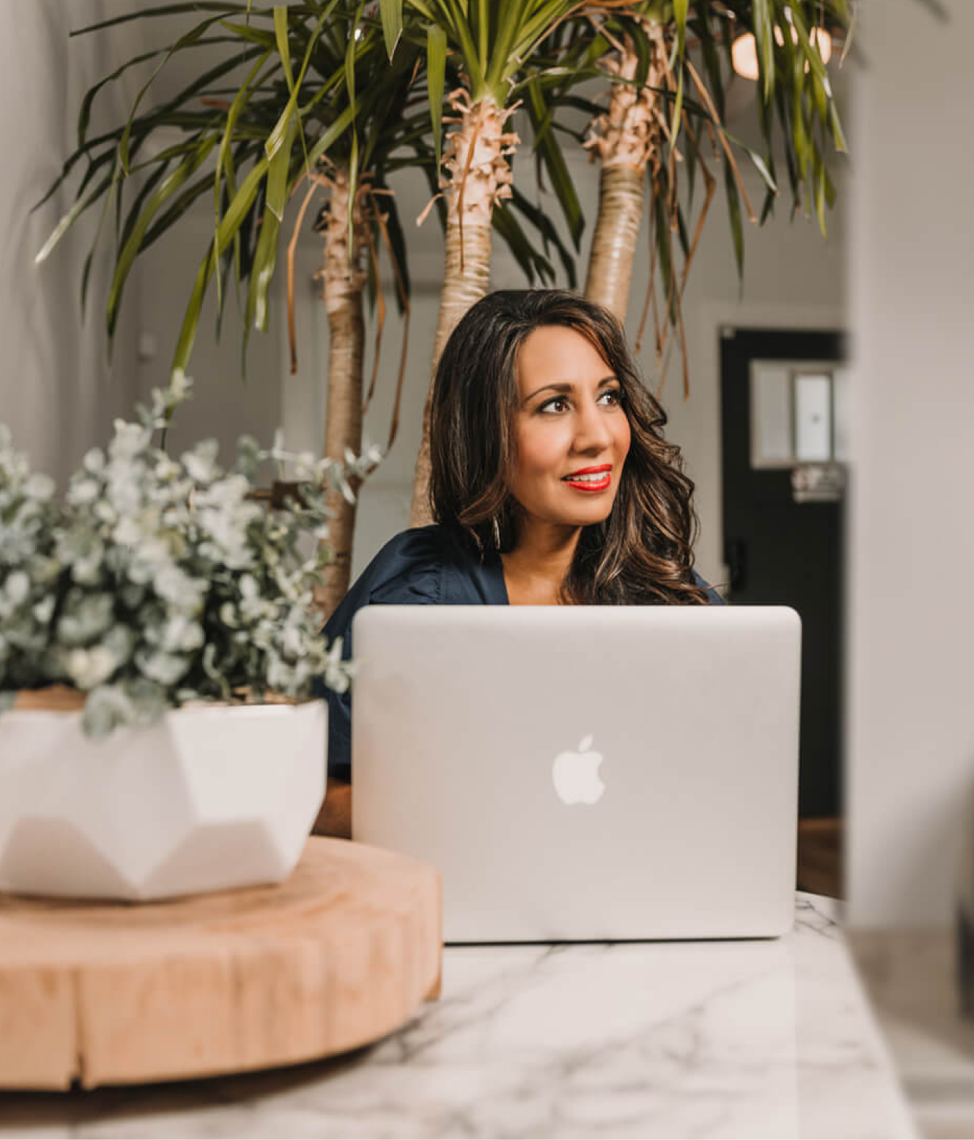 A woman with long dark hair and red lipstick sits at a marble table, looking at a laptop while surrounded by indoor plants.