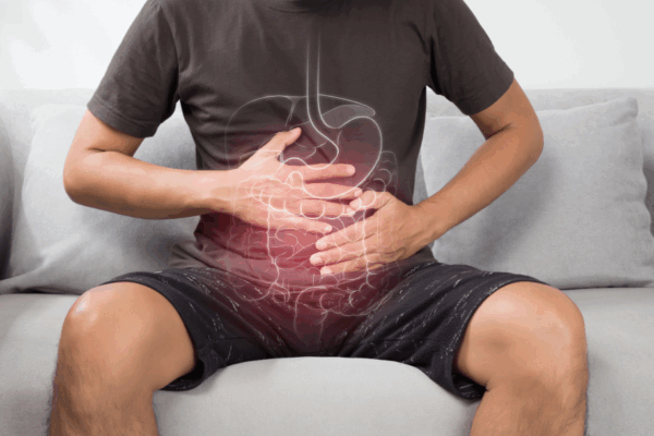 A man in a dark t-shirt and shorts sits on a couch with his hands placed on his abdomen, which is shown with a glowing anatomical illustration of digestive organs.