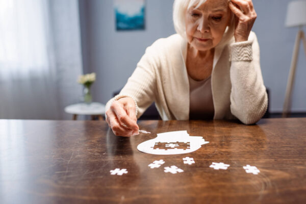 An older woman sits at a table, thoughtfully holding a puzzle piece, reflecting on Alzheimer's awareness and its impact on women.