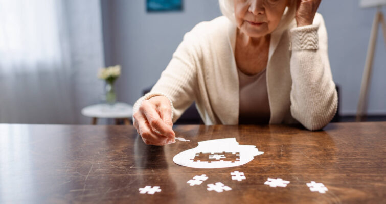 An older woman sits at a table, thoughtfully holding a puzzle piece, reflecting on Alzheimer's awareness and its impact on women.