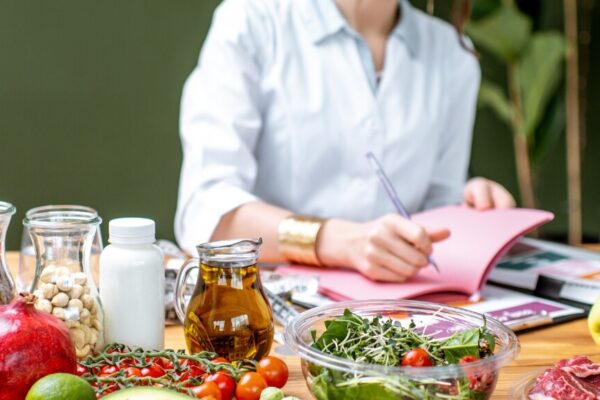 A person in a light blue shirt is sitting at a table covered with various fruits, vegetables, and ingredients while writing in a pink notebook.