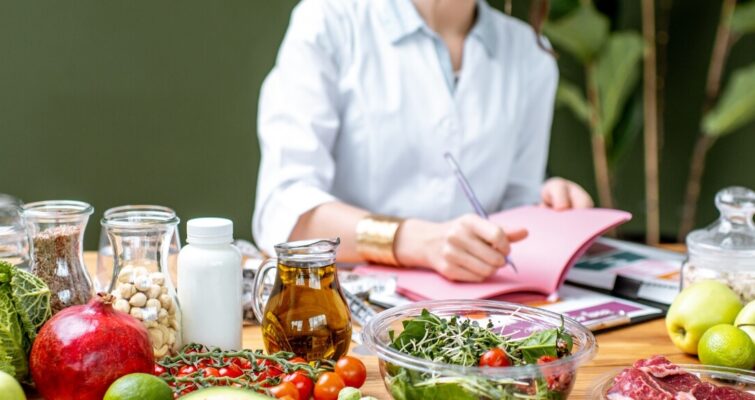 A woman sits at a table surrounded by an array of fresh fruits and vegetables, showcasing a vibrant, healthy spread.