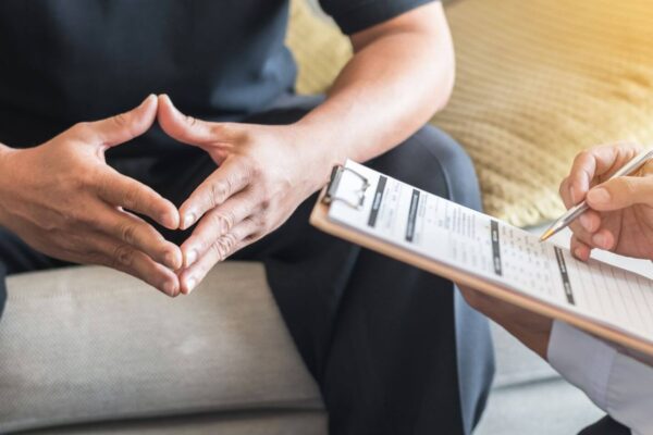 A man sits on a couch with a clipboard while a woman speaks to him, engaged in a conversation.