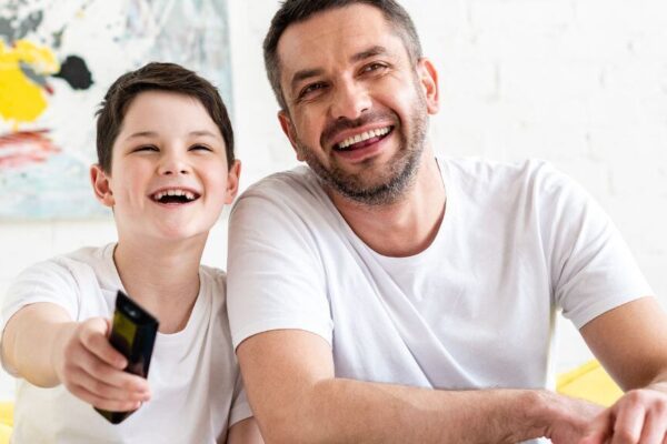 A man and a boy sit on a couch, holding a remote control, engaged in a relaxed moment together.