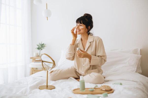 A woman sits on a bed, holding a toothbrush, with a mirror reflecting her focus on skincare and beauty trends.