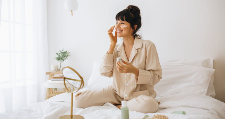 A woman sits on a bed, holding a toothbrush, with a mirror reflecting her focus on skincare and beauty trends.