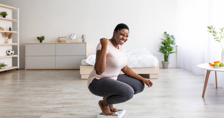 A woman squats on a scale in her bedroom, checking her weight while surrounded by soft lighting and personal items.
