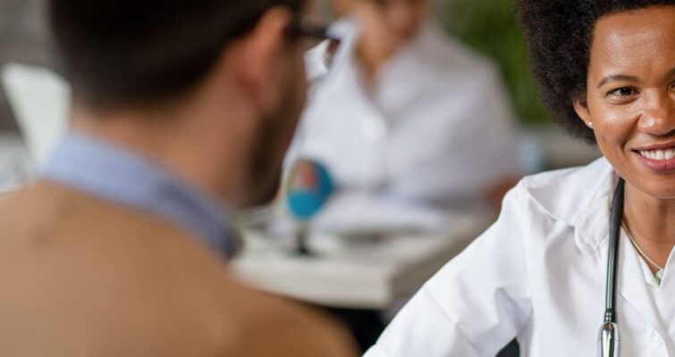 A woman in a white coat discusses with a man, likely about integrative primary care versus consulting practices.