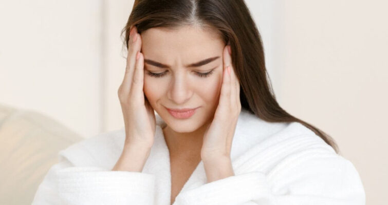 A woman in a bathrobe holds her head, possibly indicating discomfort related to mono symptoms.