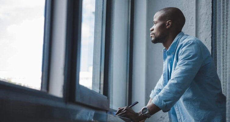 A man wearing a light blue shirt stands by a window, looking thoughtfully outside while holding a pen and notebook.