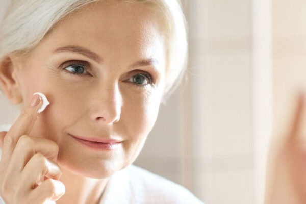 A woman in her 40s applies cream to her face while looking in the mirror, focusing on her skincare routine.