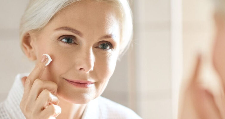 A woman in her 40s applies cream to her face while looking in the mirror, focusing on her skincare routine.