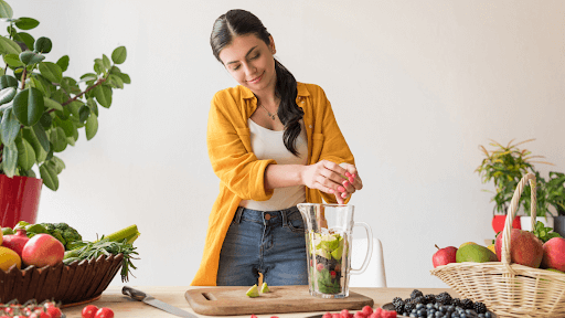 A woman blends a smoothie with fresh fruits and vegetables on a table, illustrating healthy detox options for weight loss.