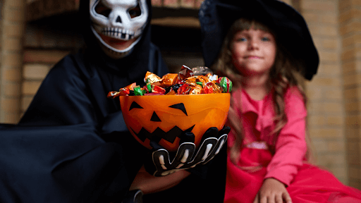 A little girl in a Halloween costume joyfully holds a bowl of candy, ready for the Halloween Candy Battle.