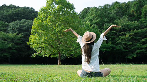 A woman sitting in grass with arms outstretched, symbolizing openness and connection to nature, related to mental health benefits.