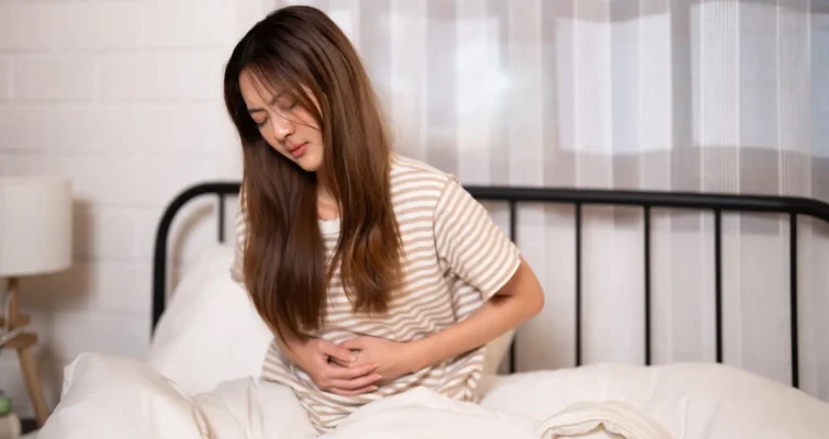 A woman sitting on a bed, holding her stomach in pain, illustrating symptoms related to SIBO and IBS.