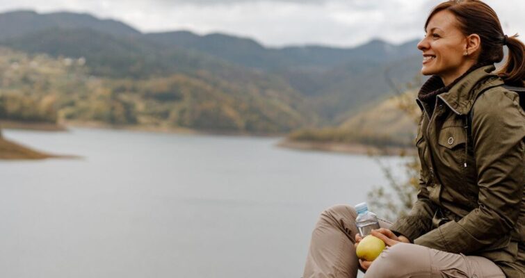 A woman sits on a hill, gazing at a serene lake, reflecting on the benefits of holistic medicine for health.