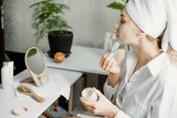 A woman in a towel applies a facial mask, highlighting a self-care moment in holistic dermatology practices.