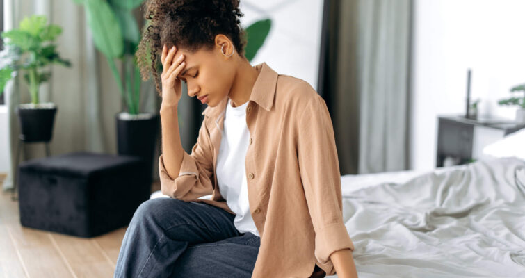 A woman sitting on a bed, holding her head in her hands, reflecting the struggle of morning migraine triggers.