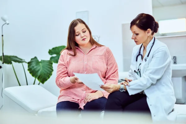A woman sitting in a doctor's office holding a paper titled 'Peptides for Weight Loss: A Functional Medicine Guide.'