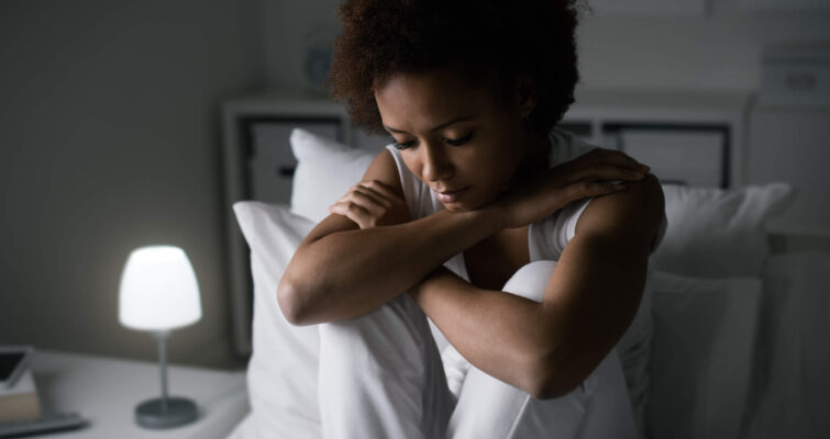 A woman sitting on a bed with her hands on her knees, reflecting on symptoms of low progesterone.