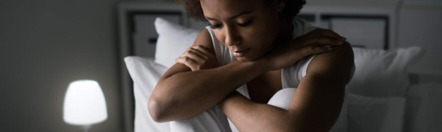 A woman sitting on a bed with her hands on her knees, reflecting on symptoms of low progesterone.
