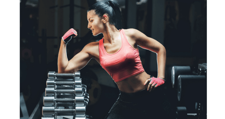 A woman in a gym poses with her arms raised, showcasing confidence and strength in a fitness environment