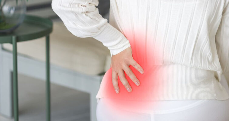 A woman sitting on a couch, visibly in discomfort, holding her lower back, illustrating back pain related to osteoporosis.