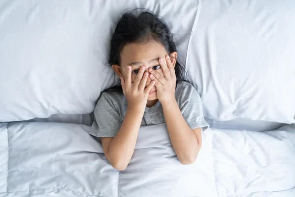 A little girl covers her eyes in bed, possibly indicating a struggle with sleep or anxiety related to bedtime.