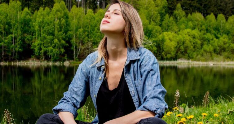 A woman sitting on the grass by a serene lake, reflecting on the concept of Ojas in Ayurveda.