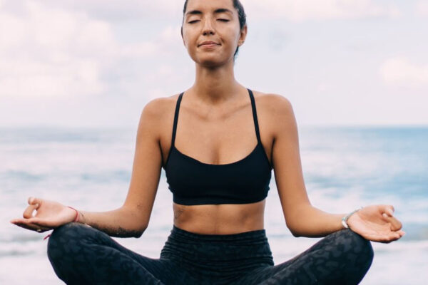 A woman in a sports bra and shorts practices yoga on the beach, surrounded by sand and ocean waves.