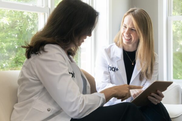 Two women in white lab coats are sitting on a couch, one smiling and pointing at a tablet while the other looks down at it.