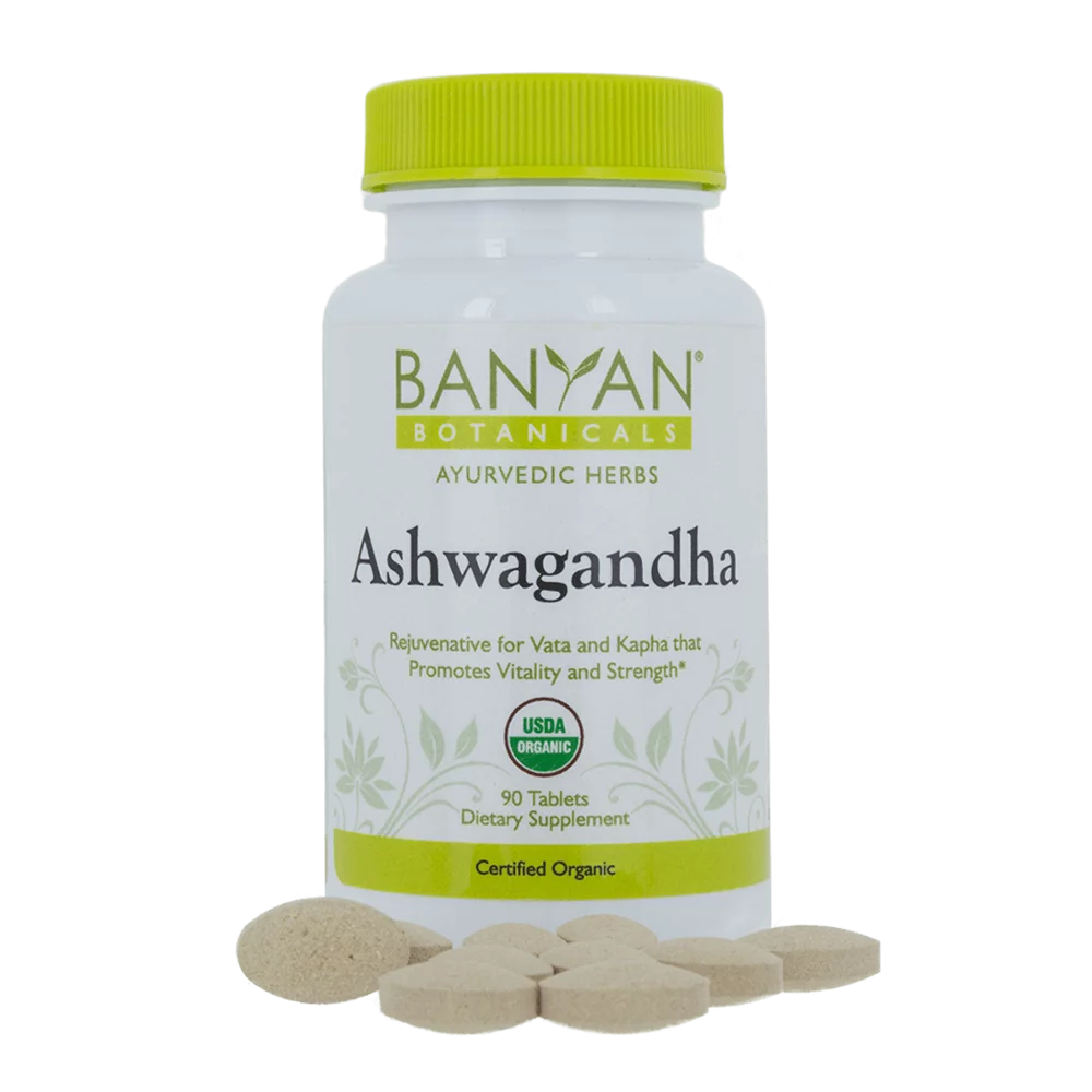A white bottle with a yellow lid labeled "Ashwagandha" contains several beige tablets arranged around it on a white background.